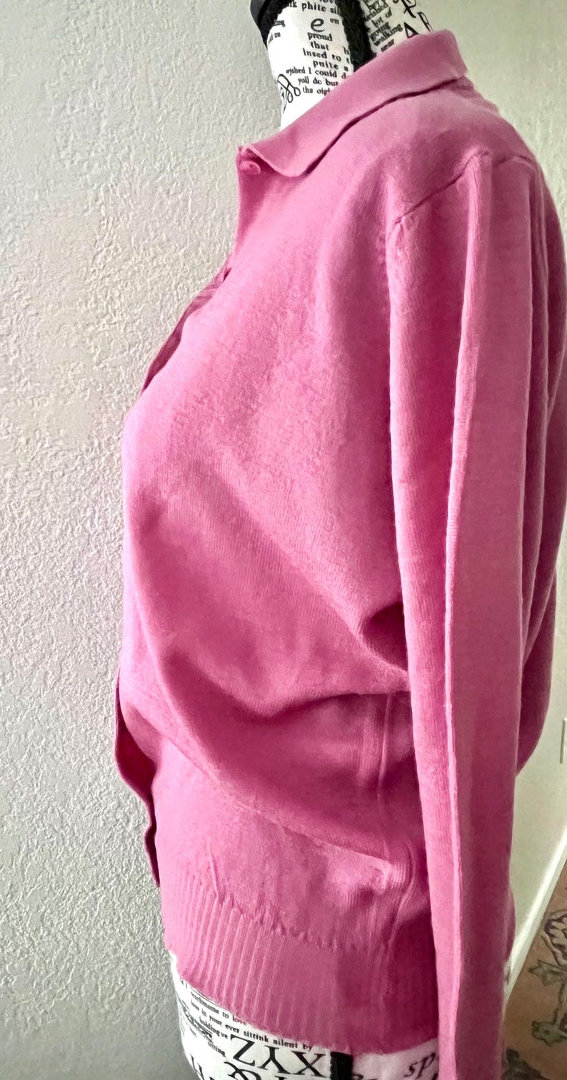 May include: A long-sleeved, solid pink knit sweater featuring a classic collar and a button-down front placket. The garment is crafted from a fine-gauge knit material and includes a ribbed hemline at the waist, displayed on a mannequin.