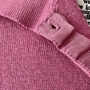 May include: A mauve-colored knit garment featuring a ribbed texture and a reinforced buttonhole placket with a visible loose thread. The fabric has a consistent weave, and a portion of a white label with black printed text is visible in the corner.