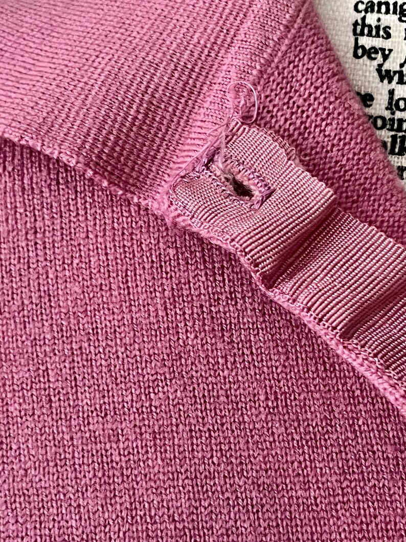May include: A mauve-colored knit garment featuring a ribbed texture and a reinforced buttonhole placket with a visible loose thread. The fabric has a consistent weave, and a portion of a white label with black printed text is visible in the corner.