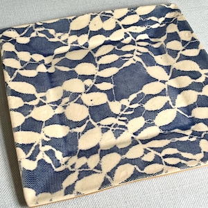 Puede incluir: Azulejo cerámico cuadrado con fondo azul y un patrón de hojas color crema. El diseño de hojas recuerda al encaje, creando una apariencia delicada y texturizada. El azulejo tiene un borde marrón claro. Dimensiones: aproximadamente 20 cm x 20 cm.