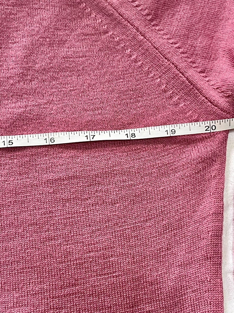 May include: A dusty rose knit sweater featuring a fine-gauge texture and a visible seam along the shoulder. A white measuring tape is positioned horizontally across the fabric, showing a measurement spanning from 15 to 20 inches in length.