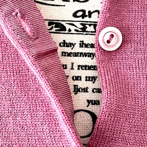 May include: A mauve knit cardigan featuring a ribbed button placket with a single light pink button. Behind the placket, a cream-colored fabric panel displays black printed text that reads RST, an, chay ihea, meanway, I renen, on my, lost ca, yua.