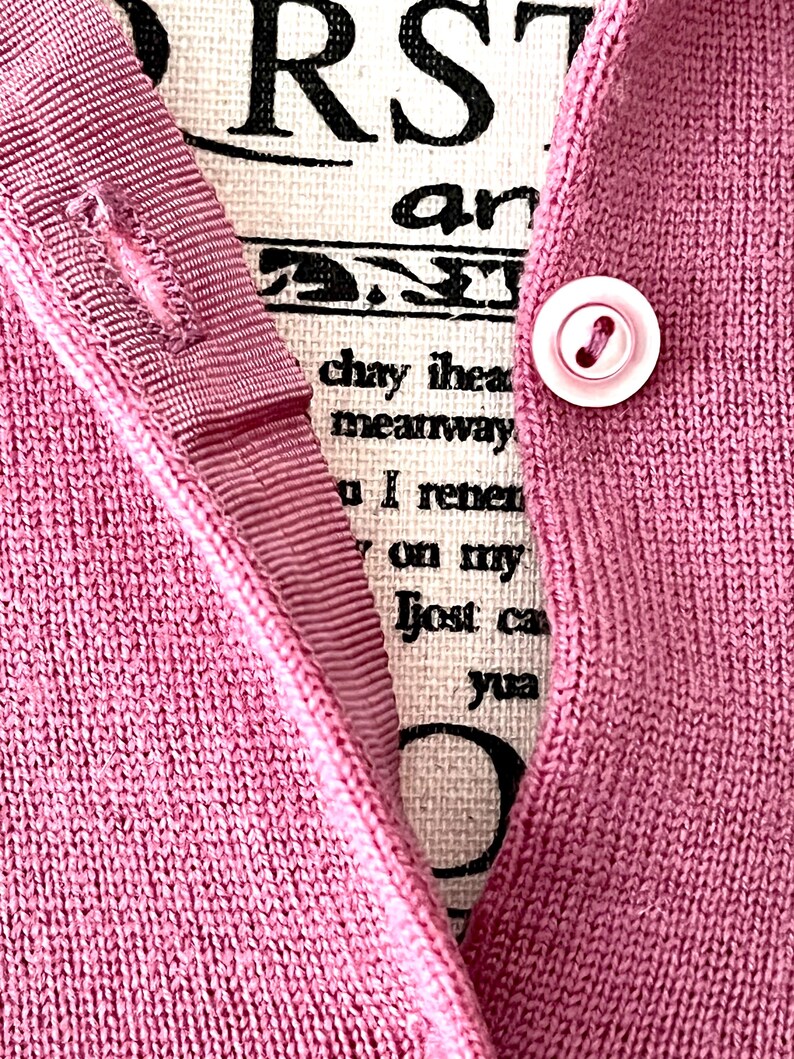 May include: A mauve knit cardigan featuring a ribbed button placket with a single light pink button. Behind the placket, a cream-colored fabric panel displays black printed text that reads RST, an, chay ihea, meanway, I renen, on my, lost ca, yua.