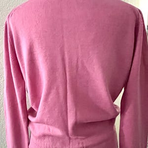 May include: A solid pink long-sleeve sweater featuring a classic collar and a ribbed hemline. The garment is displayed on a mannequin, showcasing the back view of the knit fabric, long sleeves with ribbed cuffs, and a structured collar design.
