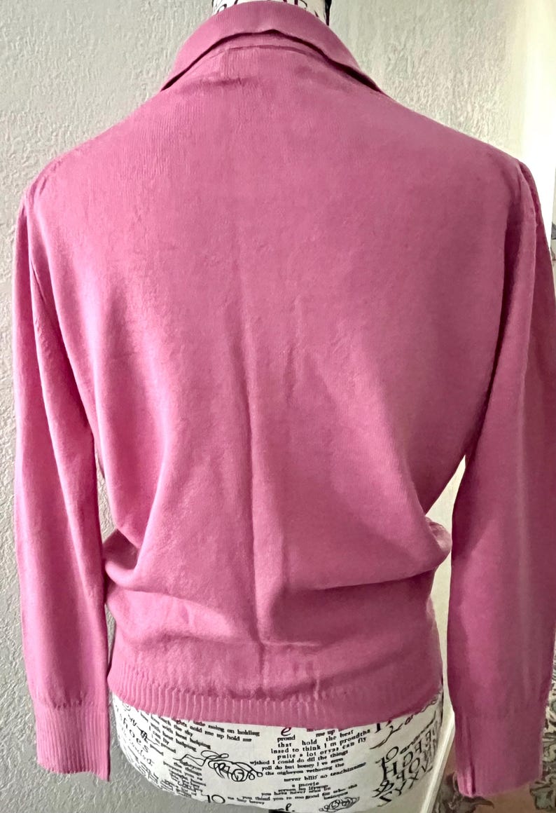 May include: A solid pink long-sleeve sweater featuring a classic collar and a ribbed hemline. The garment is displayed on a mannequin, showcasing the back view of the knit fabric, long sleeves with ribbed cuffs, and a structured collar design.