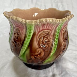 Peut inclure: Un cache-pot en céramique à bord festonné, doté d'une glaçure brillante dans des tons marron, rose et vert. La surface extérieure est ornée de motifs en relief tourbillonnants et de bandes verticales texturées entourant le bol.