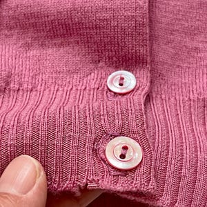 May include: A dusty rose knit cardigan featuring a ribbed texture and two round, iridescent white buttons secured with matching thread. The garment displays a fine-gauge knit construction with a vertical ribbed placket and a reinforced hemline.