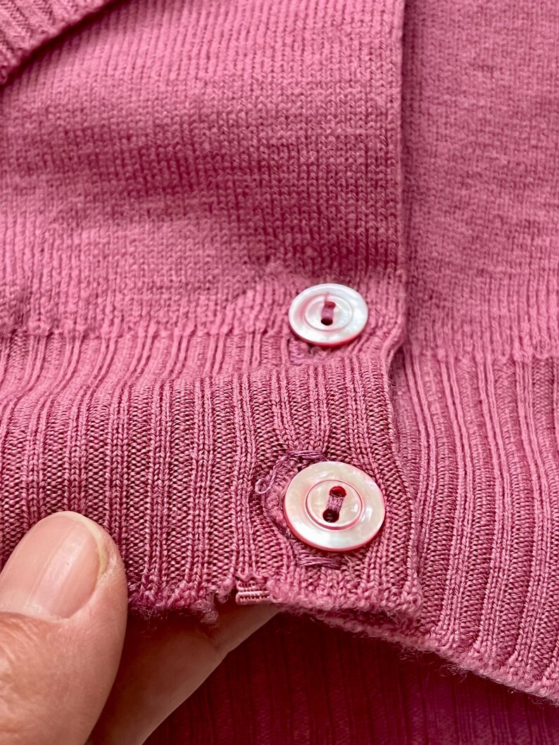 May include: A dusty rose knit cardigan featuring a ribbed texture and two round, iridescent white buttons secured with matching thread. The garment displays a fine-gauge knit construction with a vertical ribbed placket and a reinforced hemline.