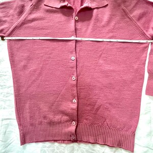 May include: A dusty rose long-sleeve cardigan with a collared neckline and a button-down front. A white measuring tape is placed horizontally across the chest, indicating a width of approximately 20 inches from side to side at the underarm area.