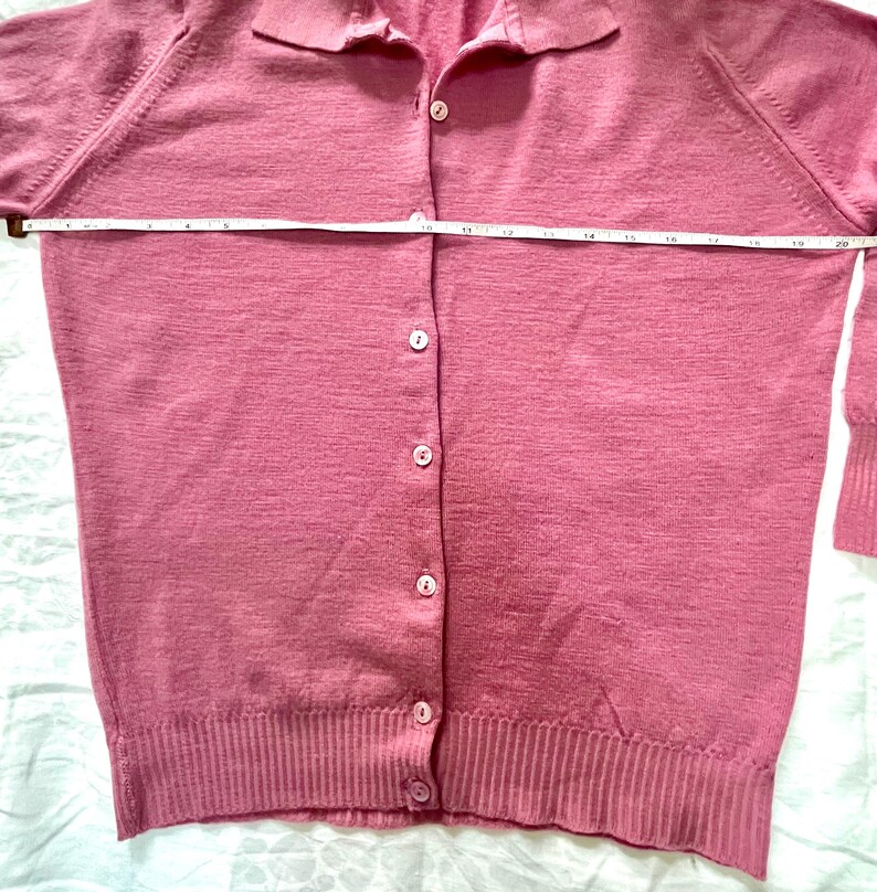May include: A dusty rose long-sleeve cardigan with a collared neckline and a button-down front. A white measuring tape is placed horizontally across the chest, indicating a width of approximately 20 inches from side to side at the underarm area.