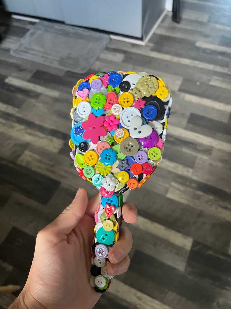 Peut inclure: Une brosse &agrave; cheveux color&eacute;e d&eacute;cor&eacute;e de boutons de diff&eacute;rentes formes, tailles et couleurs. Les boutons sont dispos&eacute;s de mani&egrave;re al&eacute;atoire, cr&eacute;ant un design unique et accrocheur.