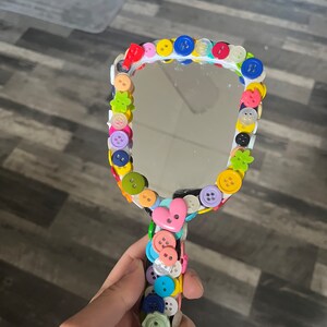 Peut inclure: Un miroir de poche blanc avec un bouton en forme de c&oelig;ur sur la poign&eacute;e. Le miroir est d&eacute;cor&eacute; de boutons color&eacute;s de diff&eacute;rentes formes et tailles, notamment des fleurs, des &eacute;toiles et des cercles.