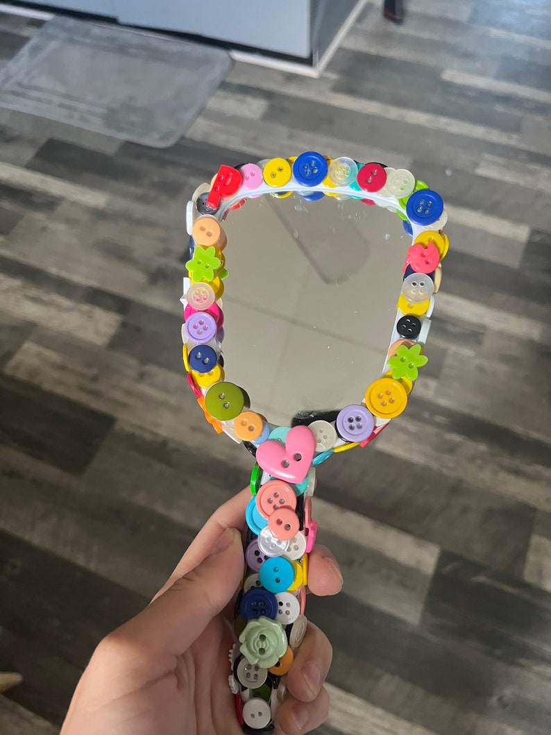 Peut inclure: Un miroir de poche blanc avec un bouton en forme de c&oelig;ur sur la poign&eacute;e. Le miroir est d&eacute;cor&eacute; de boutons color&eacute;s de diff&eacute;rentes formes et tailles, notamment des fleurs, des &eacute;toiles et des cercles.