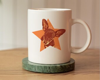 May include: A white ceramic mug featuring a graphic of a small brown dog with large ears centered inside an orange five-pointed star. The mug has a smooth cylindrical body and a curved handle on the right side, providing a simple and functional design.