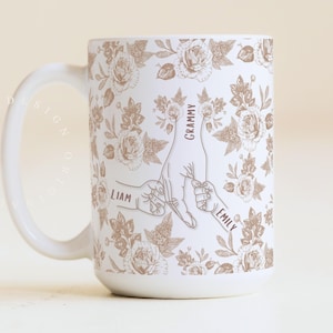 May include: White ceramic mug with a floral pattern and a design featuring hands holding each other. The names "Liam" and "Emily" are printed on the hands, with "Grammy" above. The mug has a large handle.