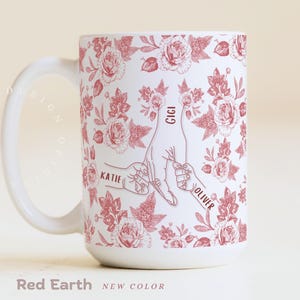 May include: White ceramic mug with a red floral pattern and the words "Katie", "Gigi", and "Oliver" on it. The mug features a design of hands holding fingers. The text "Red Earth" and "NEW COLOR" are at the bottom.