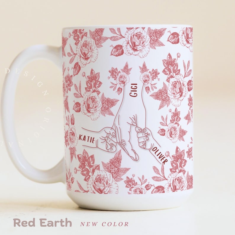 May include: White ceramic mug with a red floral pattern and the words "Katie", "Gigi", and "Oliver" on it. The mug features a design of hands holding fingers. The text "Red Earth" and "NEW COLOR" are at the bottom.