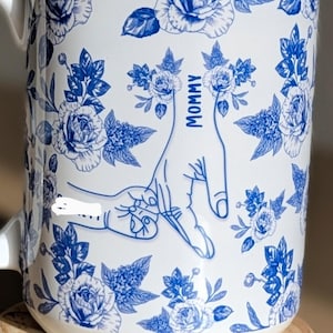 May include: White ceramic mug with blue floral design and the word "Mommy" next to a hand gesture. The mug also has the word "Blue" and a blue heart. The mug is on a wooden coaster.