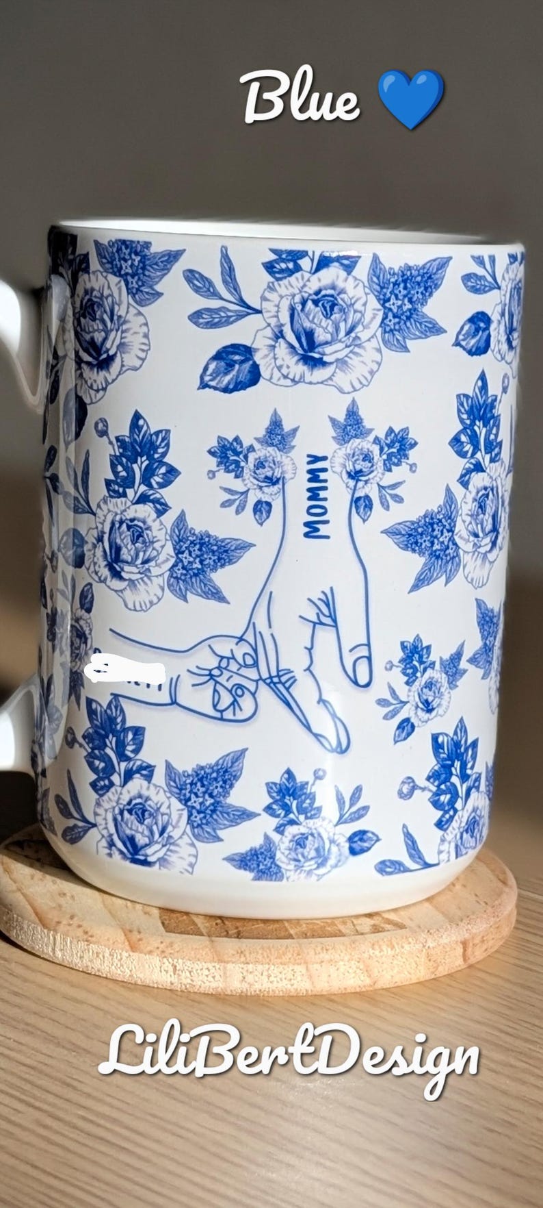 May include: White ceramic mug with blue floral design and the word "Mommy" next to a hand gesture. The mug also has the word "Blue" and a blue heart. The mug is on a wooden coaster.
