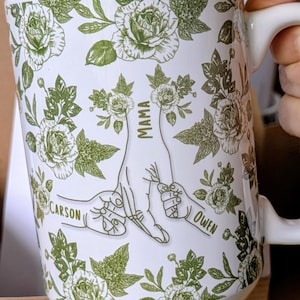 May include: White ceramic mug with a green floral pattern and the text "MAMA" with the names "CARSON" and "OWEN". The mug has a handle and is decorated with roses and leaves. The brand name "liliBertDesign" is also visible.