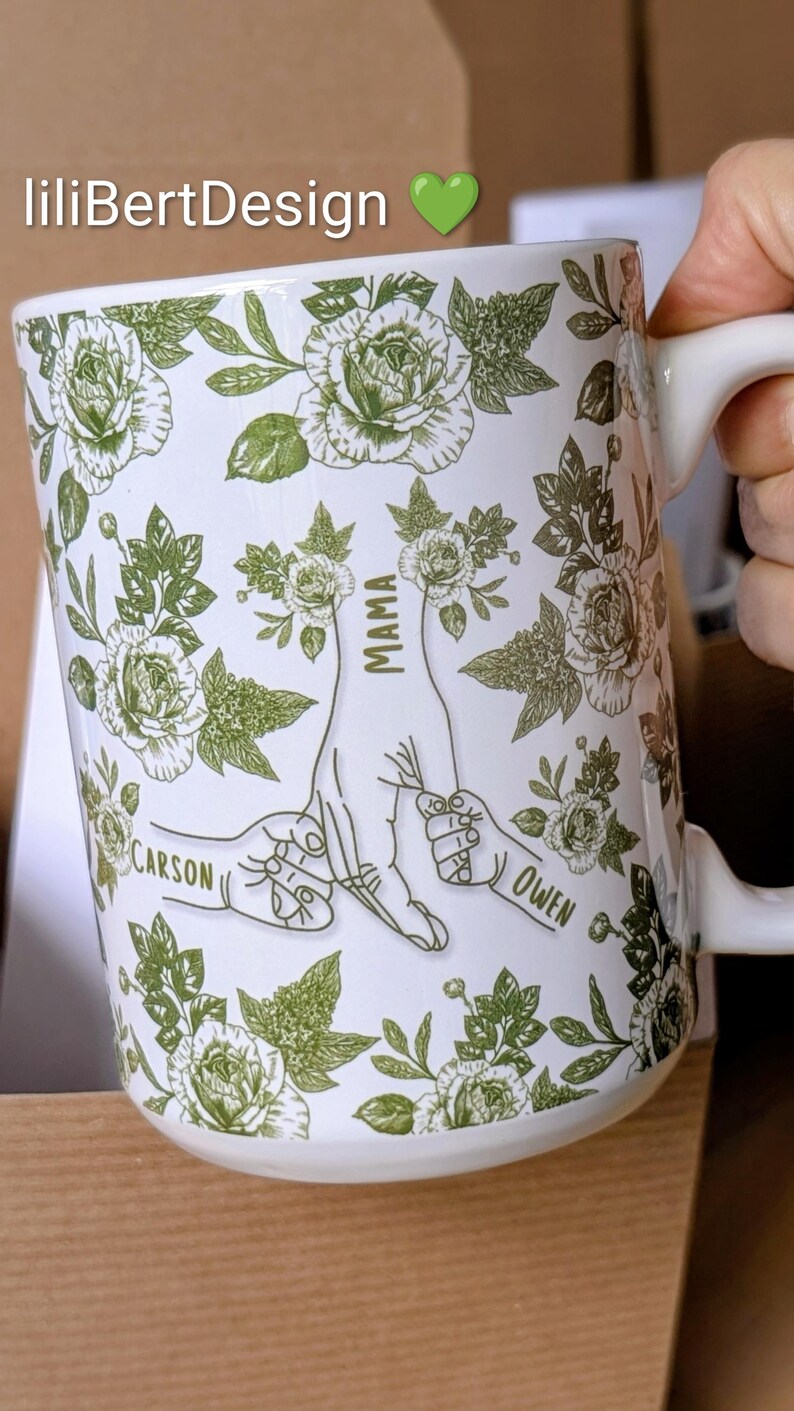 May include: White ceramic mug with a green floral pattern and the text "MAMA" with the names "CARSON" and "OWEN". The mug has a handle and is decorated with roses and leaves. The brand name "liliBertDesign" is also visible.