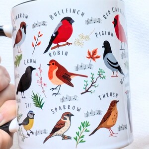 May include: White ceramic mug with a black handle and black rim. The mug features a colorful illustration of various birds, including a robin, sparrow, bullfinch, and crow. The birds are surrounded by musical notes and leaves.