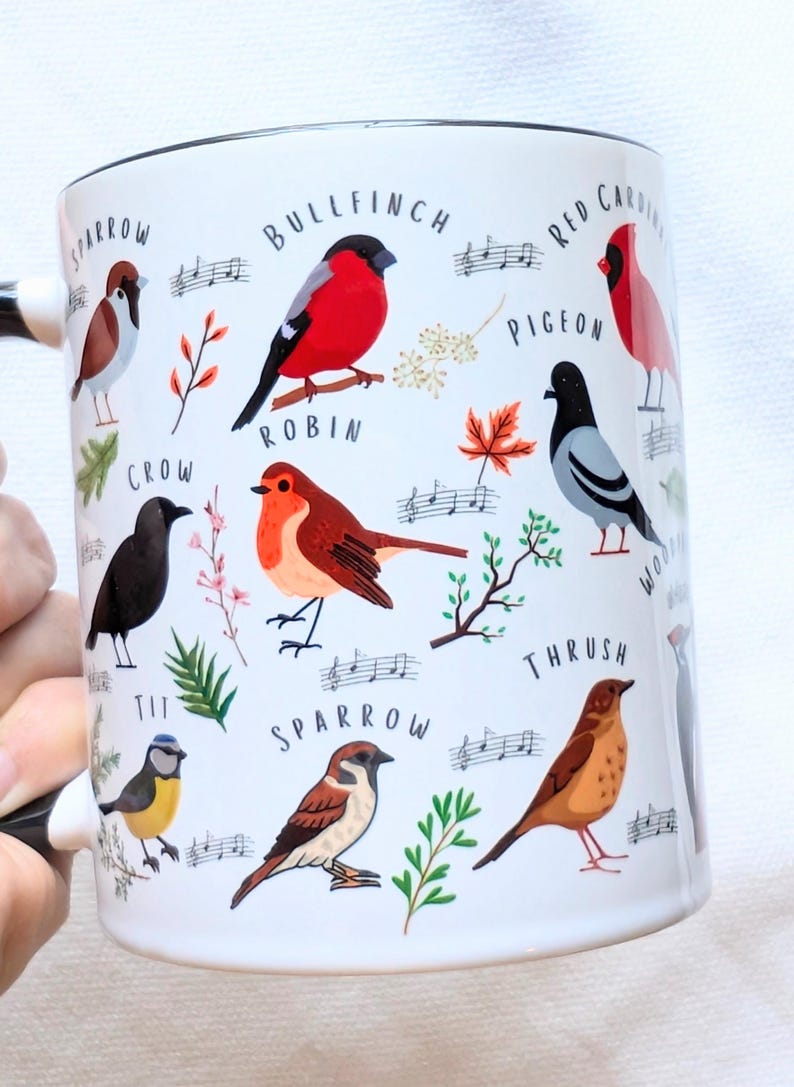 May include: White ceramic mug with a black handle and black rim. The mug features a colorful illustration of various birds, including a robin, sparrow, bullfinch, and crow. The birds are surrounded by musical notes and leaves.