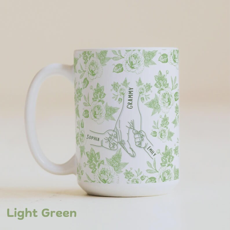 May include: White ceramic mug with a light green floral pattern and a graphic of hands holding each other. The mug has the words "Grammy", "Sophia", and "Emily" printed on it. The words "Light Green" are printed at the bottom.