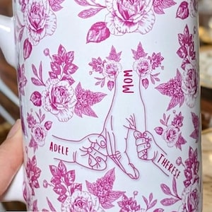 May include: White ceramic mug with a pink floral design and the words "MOM", "ADELE", and "THERESE". The mug features a pink floral pattern and a graphic of two hands holding each other. The text "liliBertDesign" is at the top.