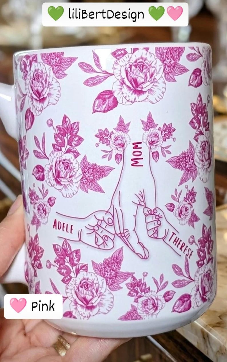 May include: White ceramic mug with a pink floral design and the words "MOM", "ADELE", and "THERESE". The mug features a pink floral pattern and a graphic of two hands holding each other. The text "liliBertDesign" is at the top.