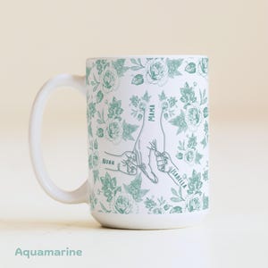 May include: White ceramic mug with a floral pattern in aquamarine. The mug features a hand-drawn illustration of hands holding each other, with the words "MAMA", "NOAH", and "ISABELLA" on the design. The word "Aquamarine" is at the bottom.
