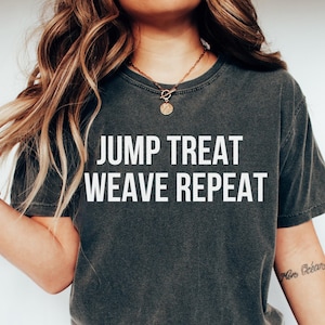 May include: Dark gray t-shirt with the text "JUMP TREAT WEAVE REPEAT" in white block letters. The shirt is a crew neck style and appears to be made of a soft, comfortable material. The shirt is worn by a person wearing blue jeans.