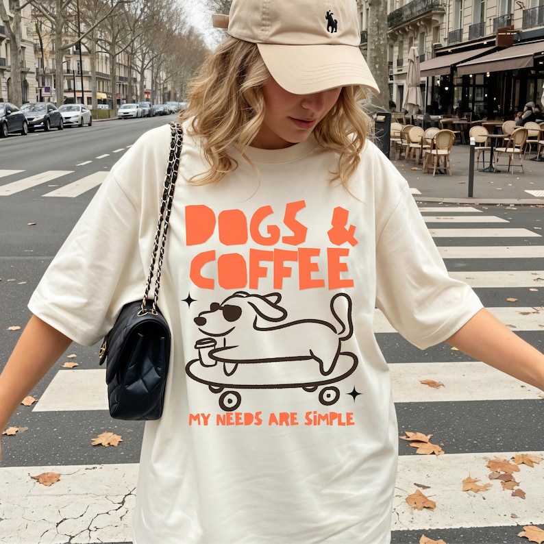 May include: An off-white short-sleeve t-shirt featuring a graphic print with the orange text "DOGS & COFFEE" above a black line drawing of a dog wearing sunglasses on a skateboard holding a coffee cup, with the orange text "MY NEEDS ARE SIMPLE" below.
