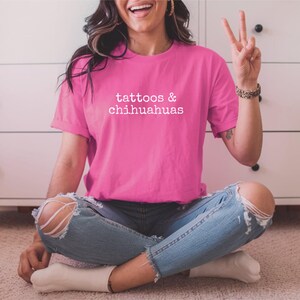 Op de afbeelding: Een vrouw draagt een roze T-shirt met de tekst "tattoos & chihuahuas" in witte letters. Ze zit met haar benen gekruist en draagt een blauwe jeans met gaten in de knie&euml;n.