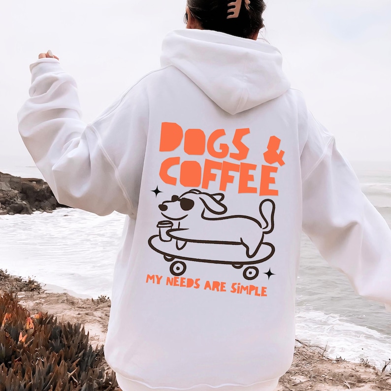 May include: A white hooded sweatshirt featuring a graphic on the back with the orange text DOGS & COFFEE above a black line drawing of a dog wearing sunglasses on a skateboard holding a cup, with the orange text MY NEEDS ARE SIMPLE below.