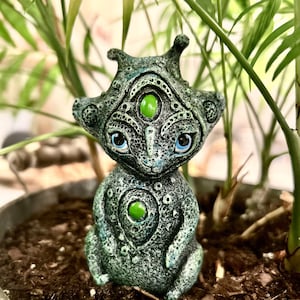 May include: A small, green, and blue ceramic figurine of a creature with three eyes and a textured surface. The creature has a rounded body and a small, pointed head.