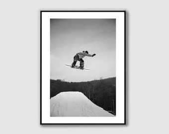 Puede incluir: Una fotografía en blanco y negro de un snowboarder en el aire, realizando un truco en una ladera nevada. El snowboarder lleva una chaqueta y pantalones, y sostiene la tabla de snowboard con la mano izquierda. El snowboarder está en el aire, y la nieve vuela a su alrededor.