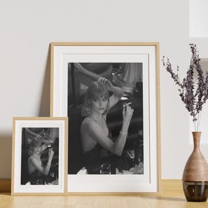 Può includere: Una fotografia in bianco e nero di una donna con i capelli corti, vestita con un abito nero, seduta su una sedia e che guarda la fotocamera. Tiene una sigaretta nella mano destra e indossa una collana. La foto è incorniciata in una cornice di legno.