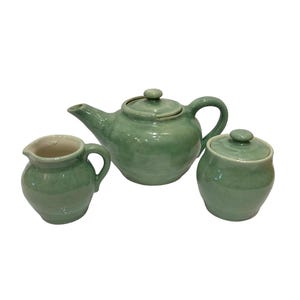 May include: A three-piece ceramic tea set in a soft green glaze. The set includes a teapot with a lid, a creamer, and a sugar bowl with a lid. The teapot has a curved spout and handle. The creamer and sugar bowl have rounded shapes.