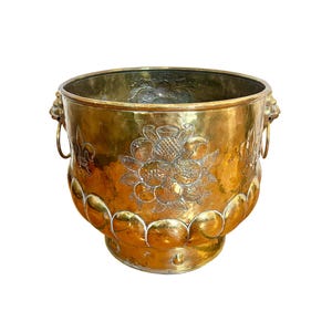 May include: A round brass planter featuring a hammered finish, lion head side handles with rings, and embossed fruit and fleur-de-lis motifs. The base is decorated with a scalloped pattern, and the metal surface displays a warm, polished gold tone.