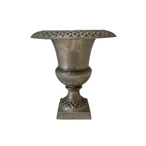 May include: A silver-colored urn with a wide, decorative rim and a flared body. The urn features intricate patterns around the base and on the square pedestal. The design suggests a vintage or antique style, suitable for home decor.