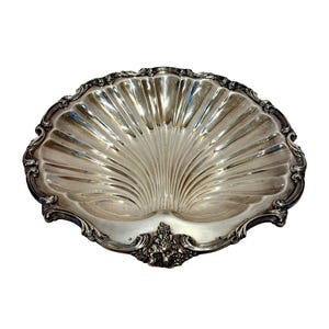 May include: A silver-colored, shell-shaped serving tray. The tray has a fluted design radiating from the center and an ornate, scrolled edge. A decorative cluster of floral elements is at the base of the shell.