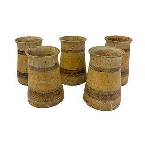 May include: A set of five ceramic tumblers featuring a speckled tan and beige glaze with horizontal bands of dark brown and muted gray. Each vessel has a flared rim, a slightly tapered cylindrical body, and a textured, earthy finish throughout.