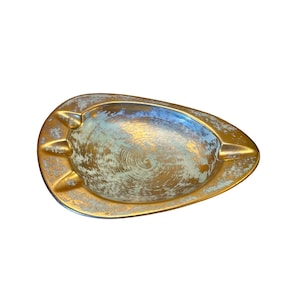 Peut inclure: Un cendrier en métal en forme de goutte d'eau avec une finition dorée et bleu clair vieillie. La surface intérieure présente une texture brossée circulaire, tandis que le large bord comporte trois encoches distinctes sur le côté gauche.