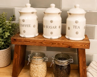 May include: Three cream-colored ceramic canisters with grey polka dots and lids, labeled COFFEE, SUGAR, and TEA, sit on a dark brown wooden riser. Below the riser are two clear glass jars with metal wire-bail lids containing dry goods.