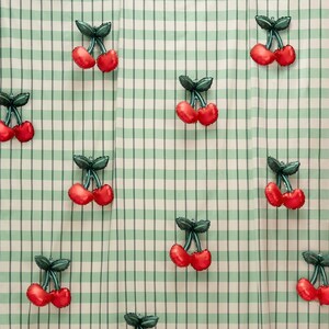 K&ouml;nnte beinhalten: Ein sich wiederholendes Muster aus roten kirschf&ouml;rmigen Luftballons mit gr&uuml;nen Stielen und Bl&auml;ttern auf einem hellgr&uuml;nen und wei&szlig;en karierten Hintergrund. Die Ballons sind &uuml;ber den Stoff verteilt und erzeugen ein fr&ouml;hliches Design.