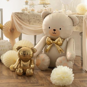 May include: Two teddy bear balloons, one large and cream-colored with a gold bow tie, and a smaller gold metallic bear. The scene includes party decorations like paper fans and a pom-pom, suggesting a baby shower or birthday celebration.