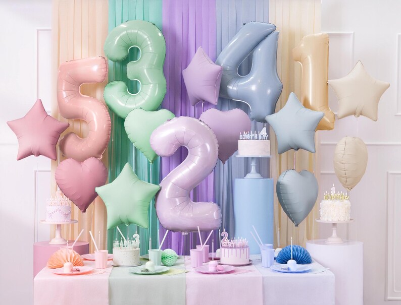 K&ouml;nnte beinhalten: Ein Geburtstagstisch im Pastellton mit Ballons in Form von Sternen und Zahlen, darunter eine gro&szlig;e Zahl 2, 3, 4, 5 und 1. Es gibt zwei Kuchen mit Kerzen, einer mit einer Krone oben drauf. Der Tisch ist mit einer rosa Tischdecke bedeckt und hat eine Vielzahl von pastellfarbenen Tellern und Tassen.