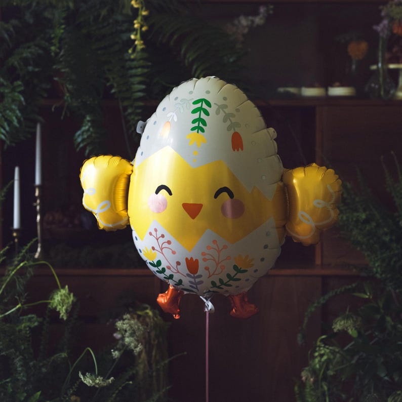 Op de afbeelding: Een vrolijke paasei-vormige ballon met een kuikendesign. De ballon heeft een witte eierschaal met bloemmotieven, gele vleugels en oranje voetjes. Het kuiken heeft een lachend gezicht met roze wangen en een kleine oranje snavel. Een roze touwtje is bevestigd.