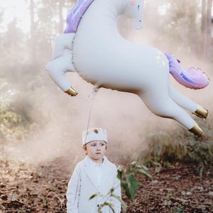May include: A young boy wearing a white crown and a white suit stands in a woodland setting with a large white unicorn balloon floating above him.